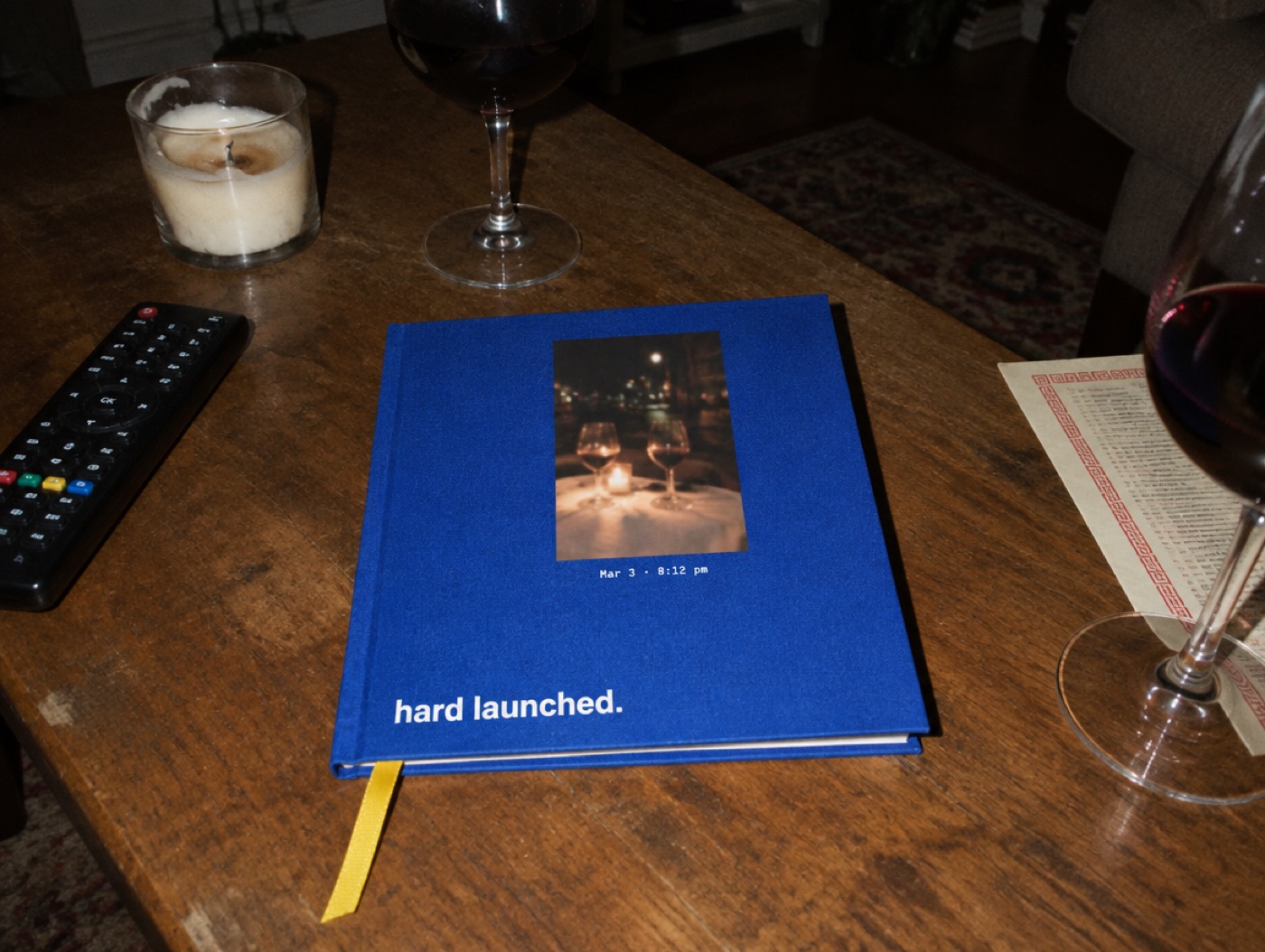 A cobalt blue hardcover on a coffee table at night. The cover has a small photo of two wine glasses, the caption Mar 3, 8:12 pm, and the title hard launched.